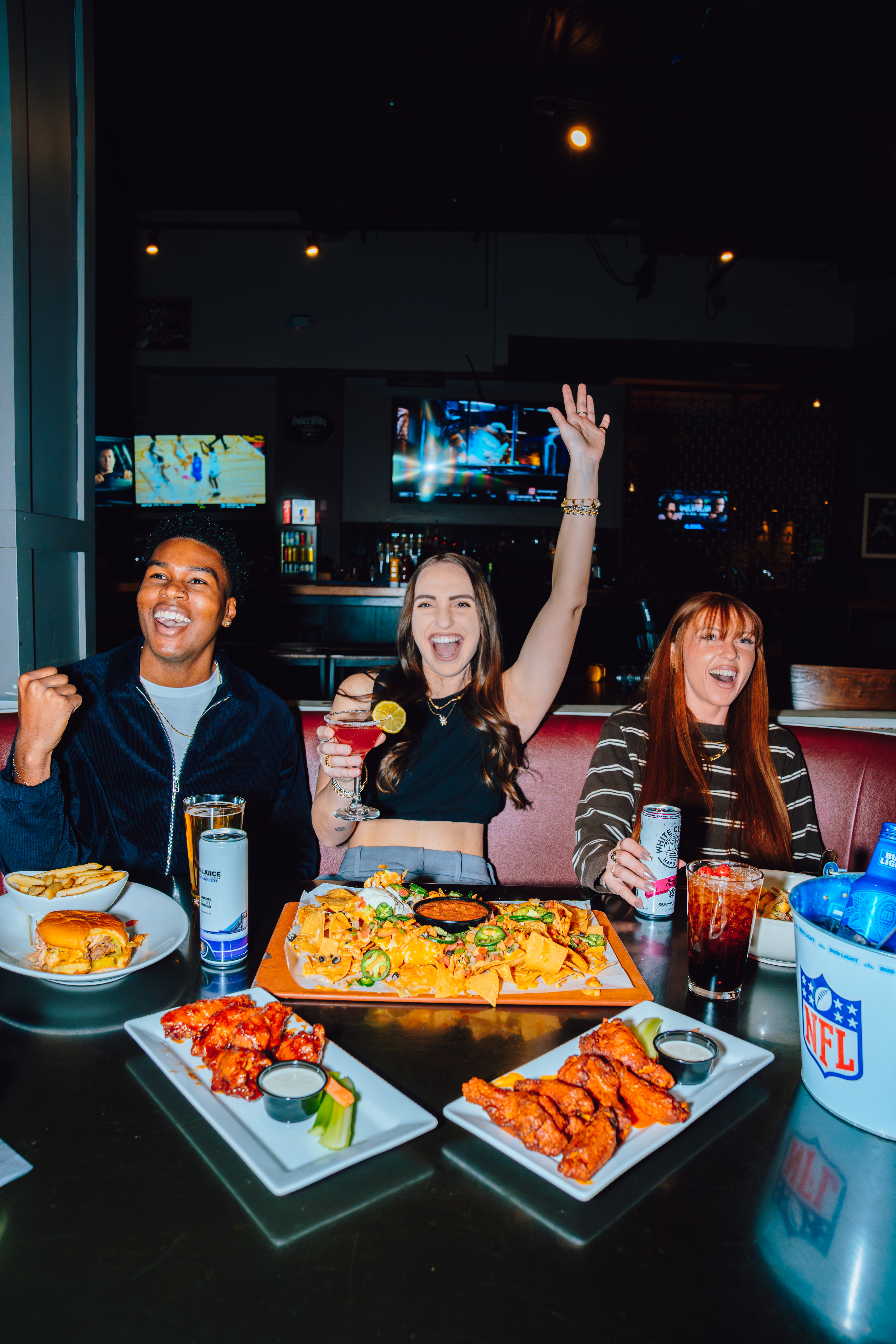 An image of 2 girls and a guy cheering at a table full of food.
