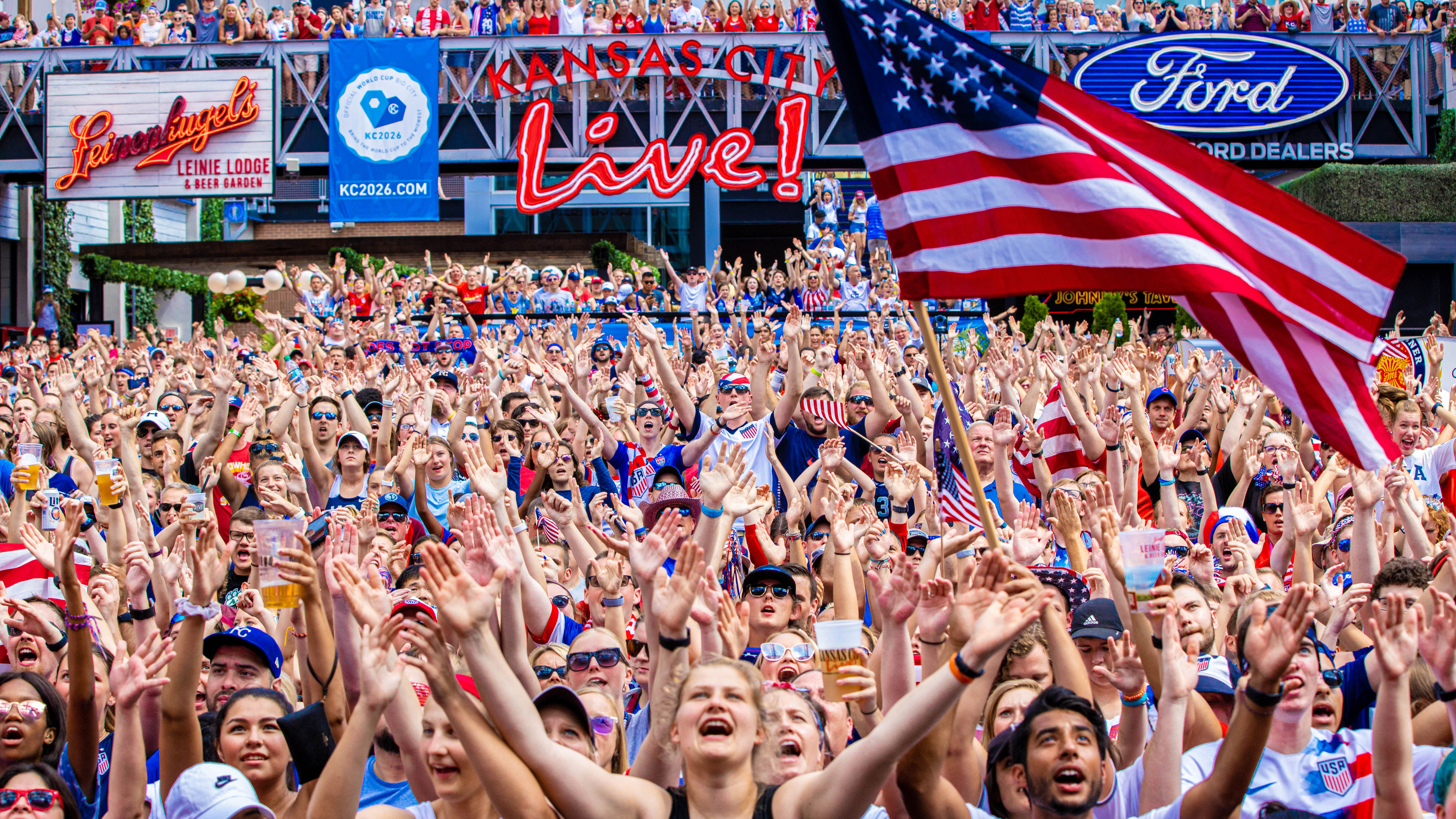 An excited crowd waves their arms and screams, with an American flag waving in the foreground and neon logos for Live! Ford, KC2026.com, and Leinenhugel's in the background.