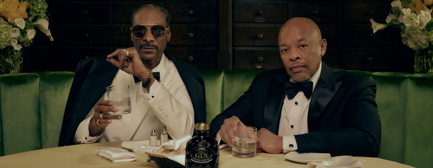 Snoop Dogg and Dr. Dre sitting in a green banquet booth with Still G.I.N. bottle on the table in front of them.