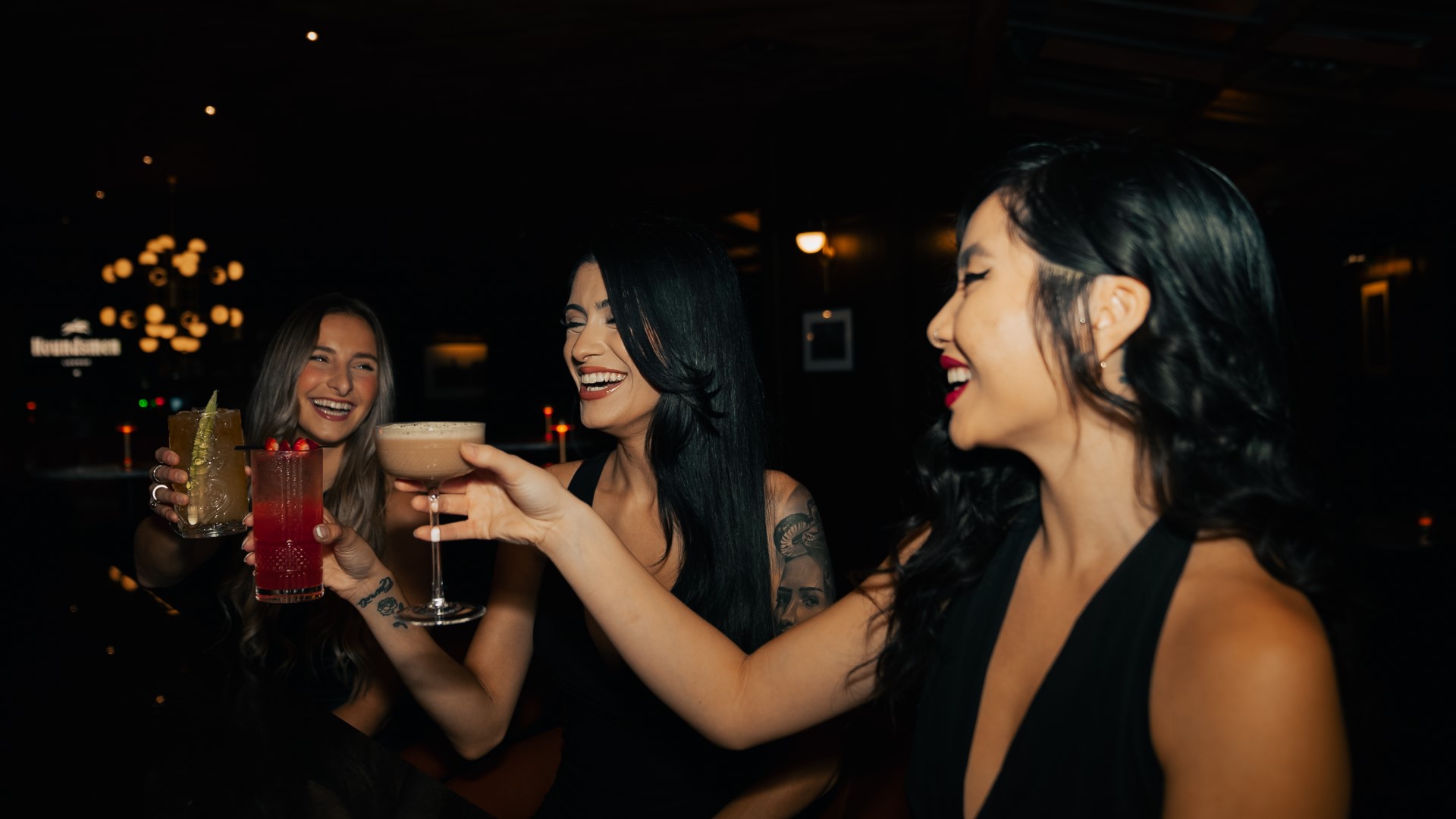 Three young women in black dresses toasting cocktails at the bar of Houndsmen Lounge in Orlando.