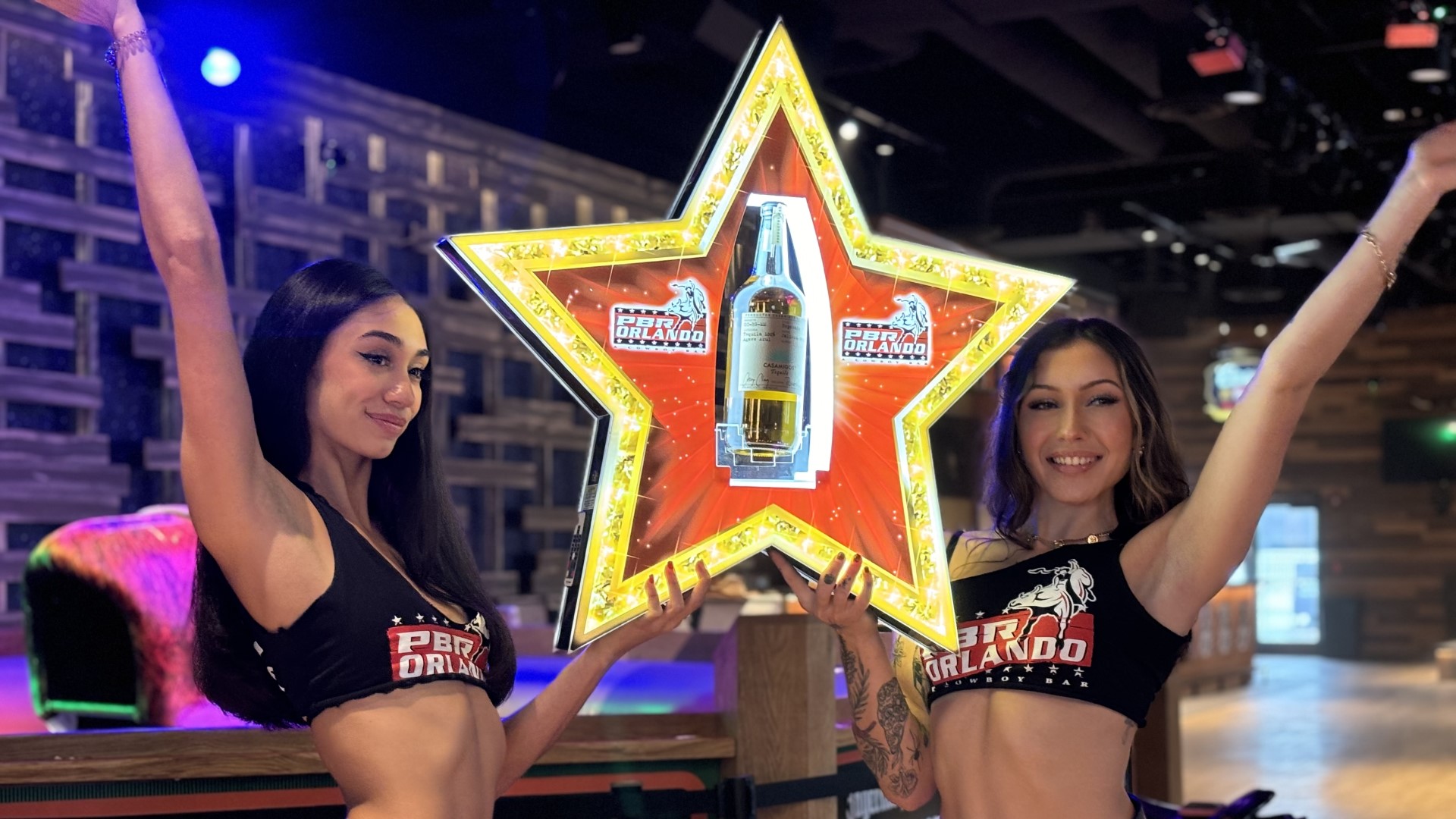Two Buckle Bunny hostesses wearing a cropped t-shirts and chaps presenting bottle service where bottle is held in a neon star sign.