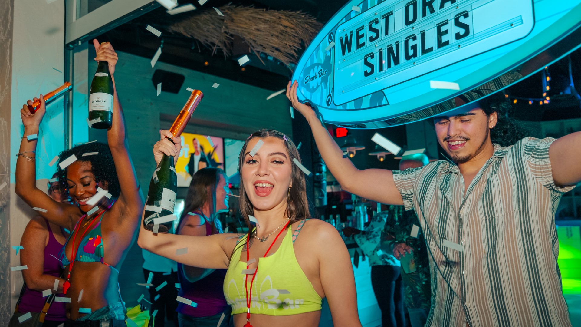 Two female and one male Shark Bar staff members presenting bottle service with confetti, surfboard sign, and light sticks.