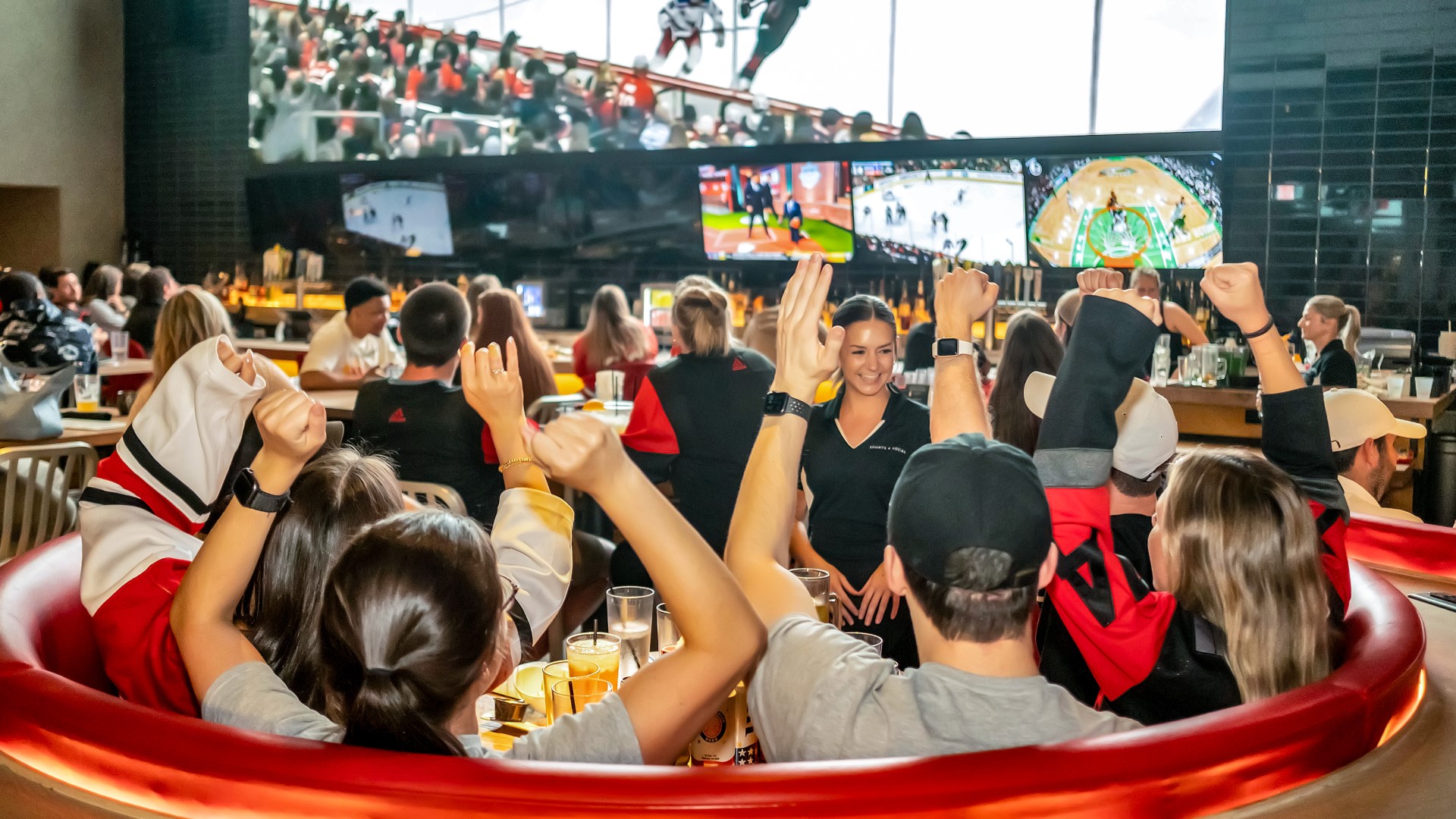 Guests at Sports & Social watching and cheering on games on big screen tvs in the bar area and booth seating.