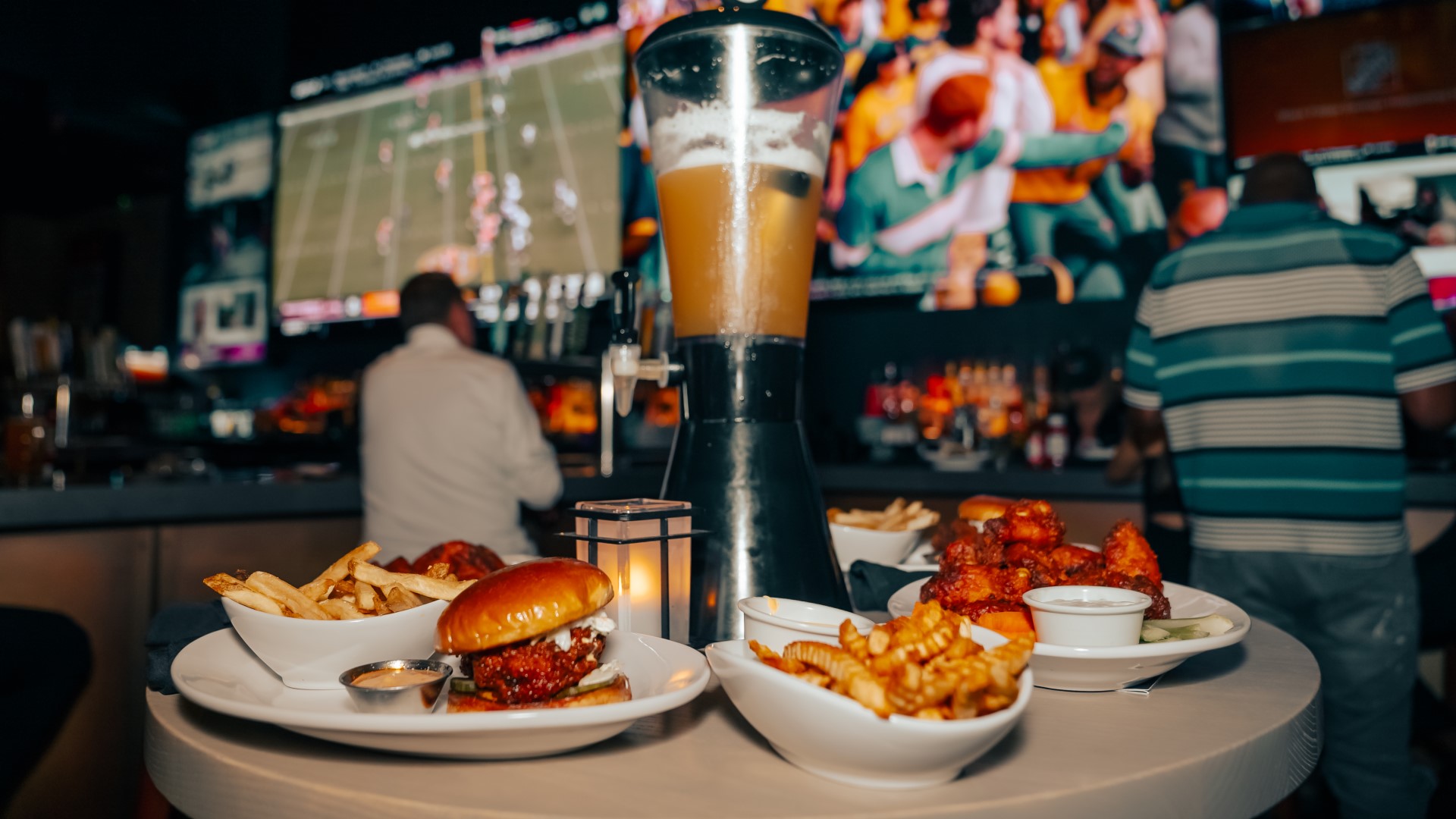 Beer tower and entrees and appetizers on white plates on a table in front of the big screen TVs and bar at Sports & Social at Live! at the Pointe Orlando.