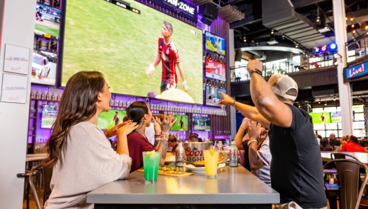 Woman and man at Sports & Social watching soccer on large screen TV and media wall.