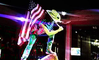 Man wearing a cowboy hat and holding an American flag standing on a mechanical bull at PBR.