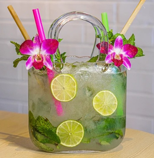 Factoria de Azucar cocktail in a purse-shaped glass with three lemon slices and mint inside and garnished with two pink tropical flowers. mint, and four straws.