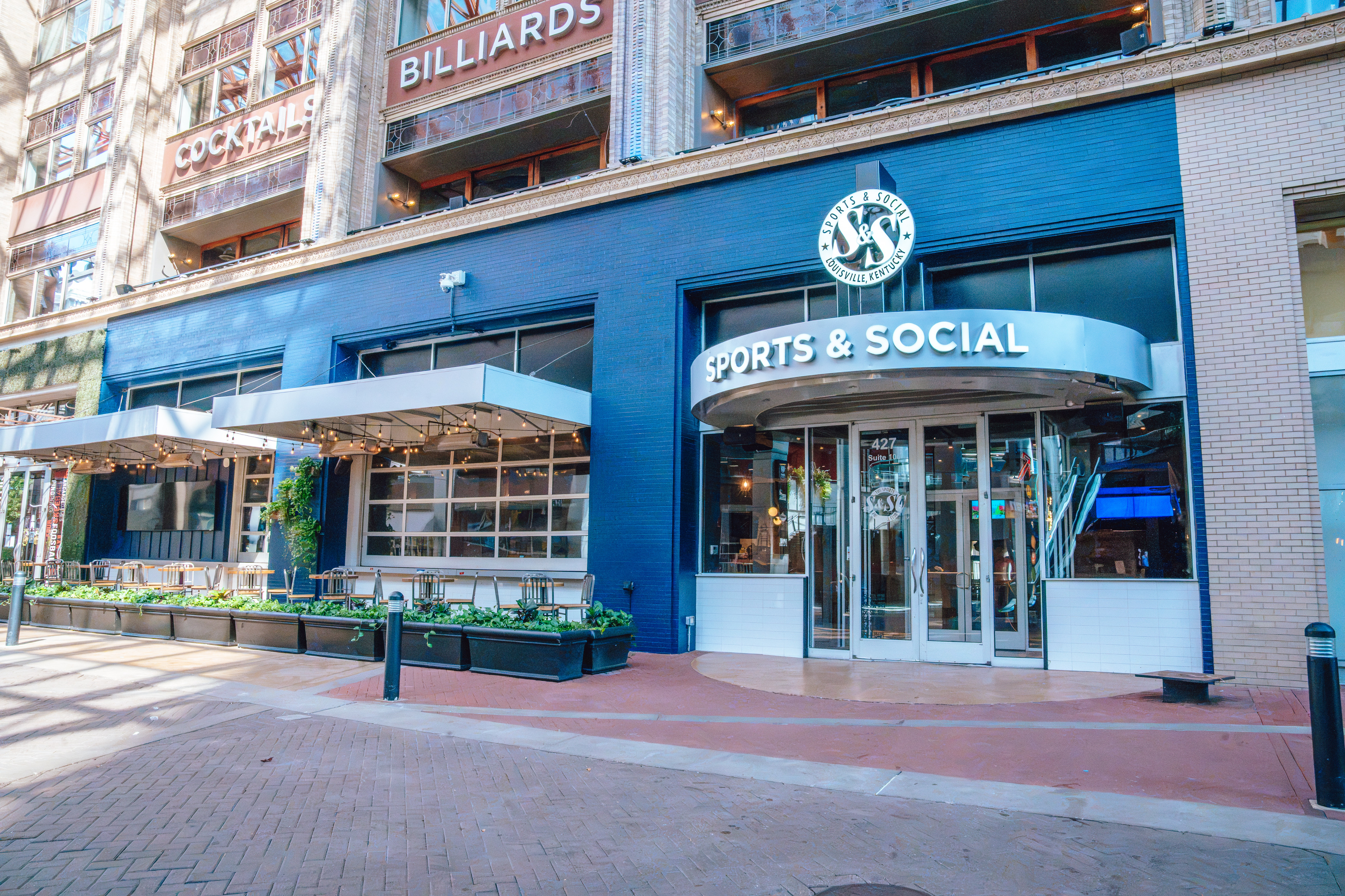 A wide shot of the exterior of Sports and Social with the new Sports and Social logo on top of the entrance and the entire patio with patio tables in view, and blue, brick walls.