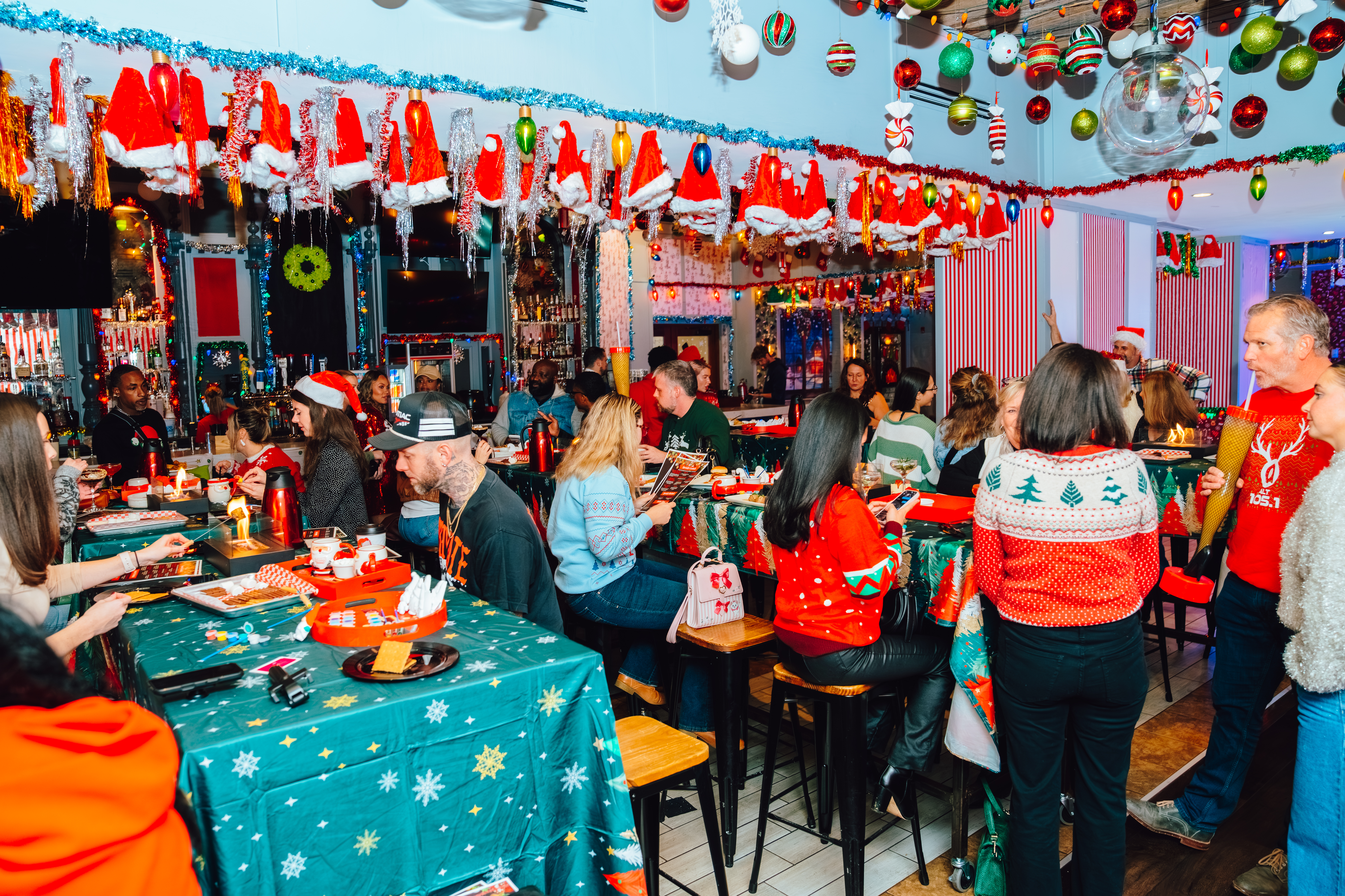 A wide shot image of the interior of Tinsel Tavern with Christmas decor covering the wall and two long tables full of people in festive red and green clothing, socializing.