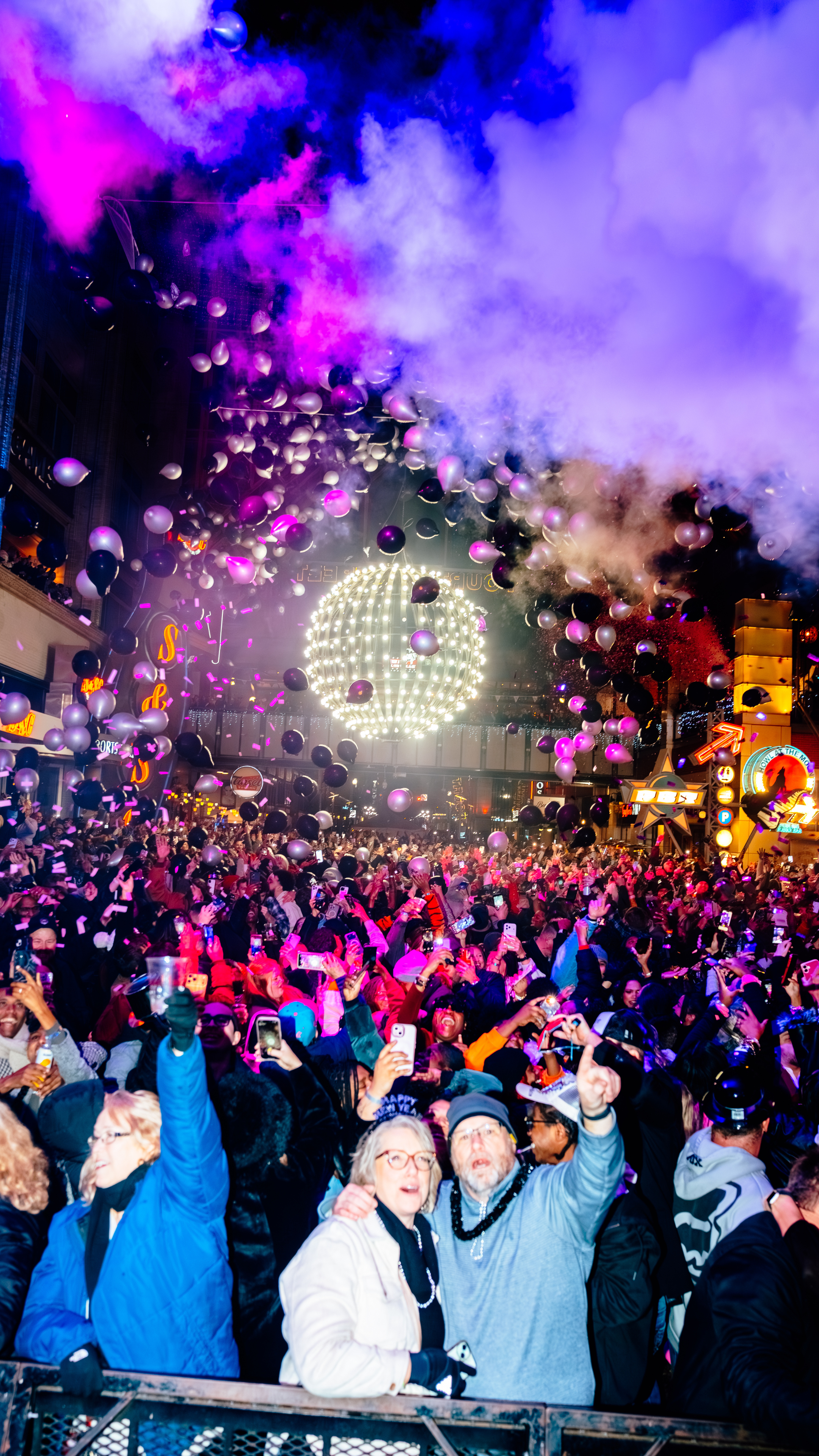 An image of the NYE ball drpping over a crowd of people, with balloons in the air and purple and pink smoke above the ball.
