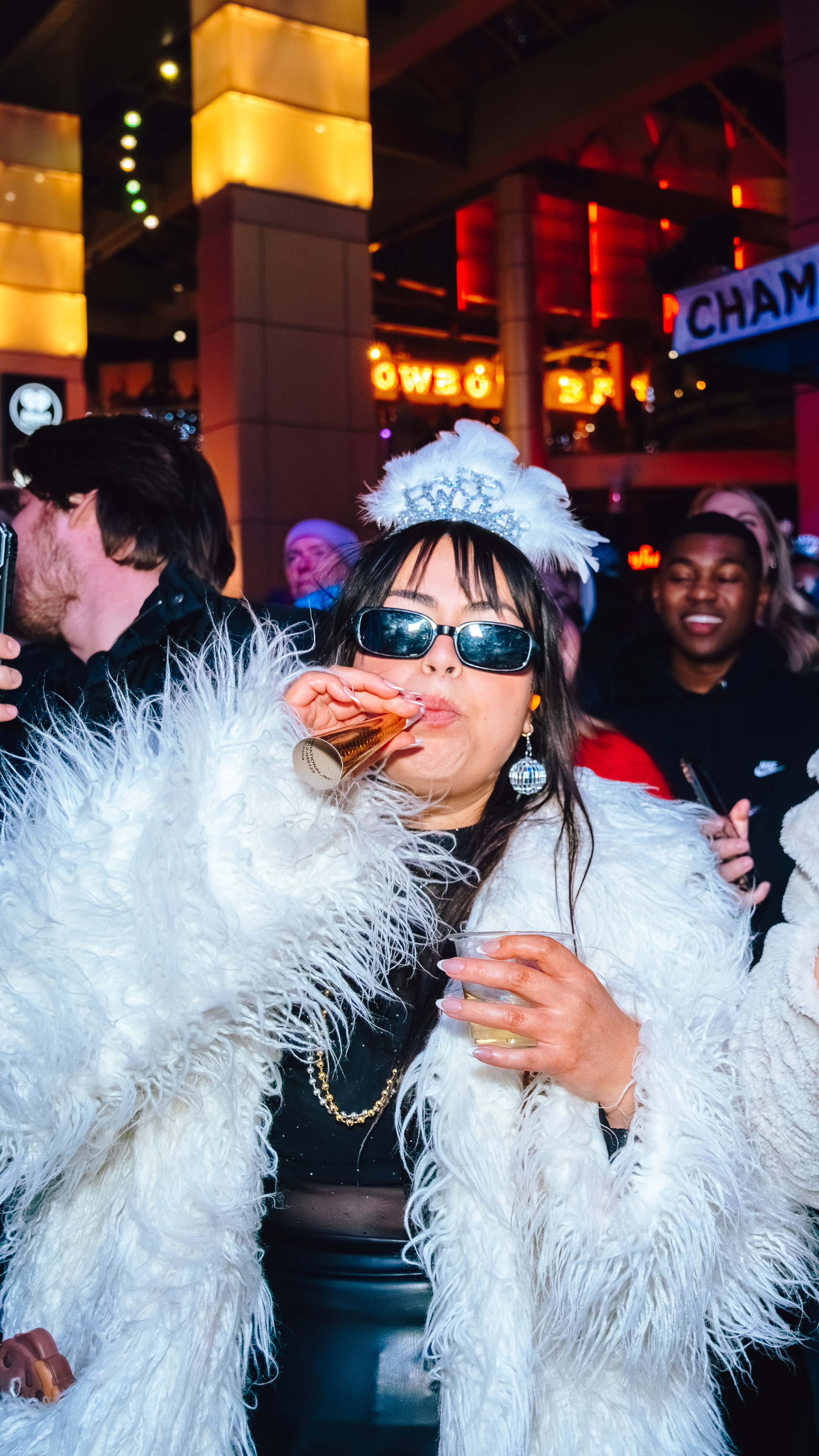 AN image of a woman wearing a white fur coat, with NYE headband and sunglasses on, and blowing into a noise maker.