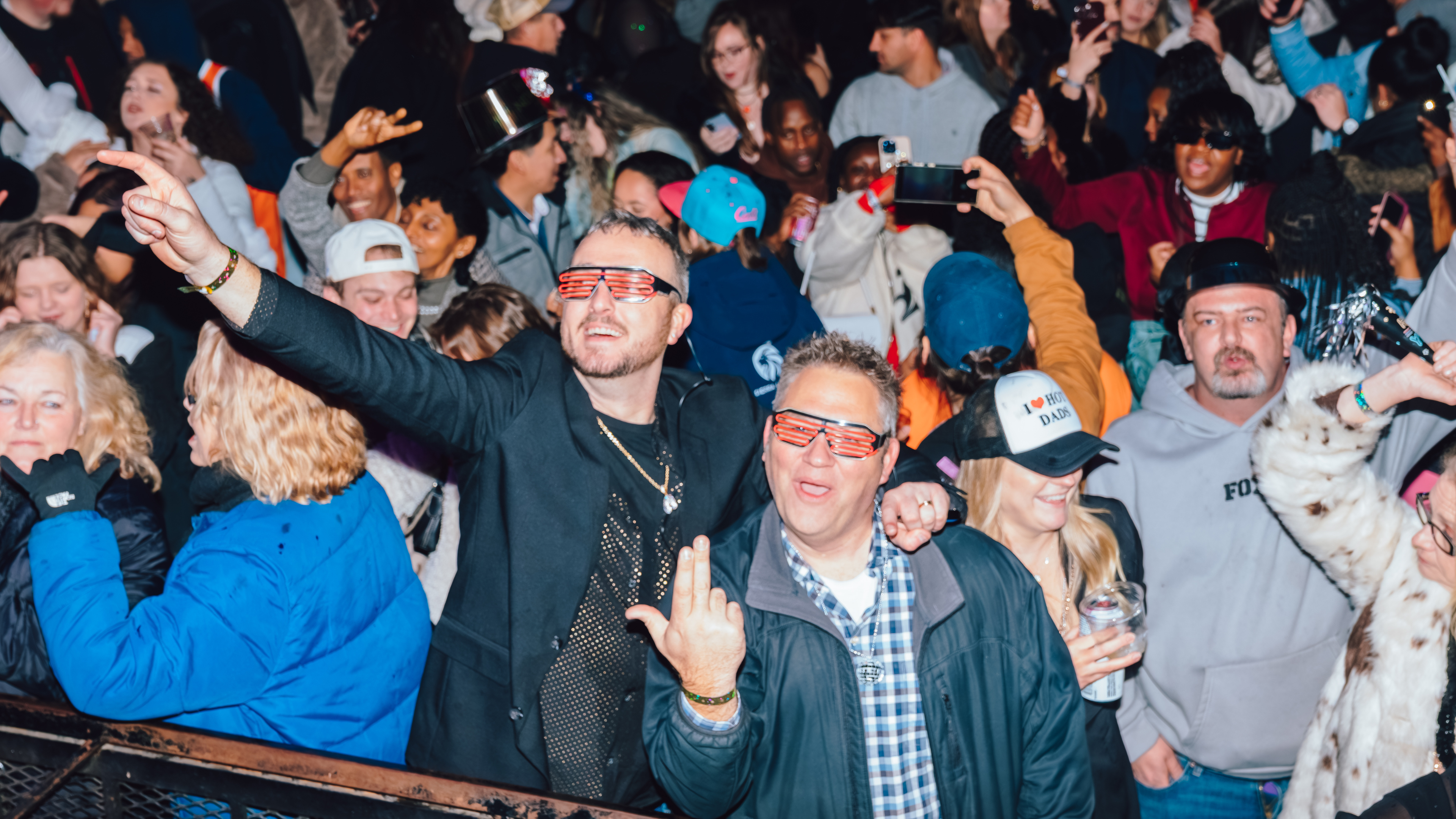 An image of two men in a crowd of people at an NYE party, with one pointing up towards the sky, and the other throwing up an "L" with his hand, for Louisville.