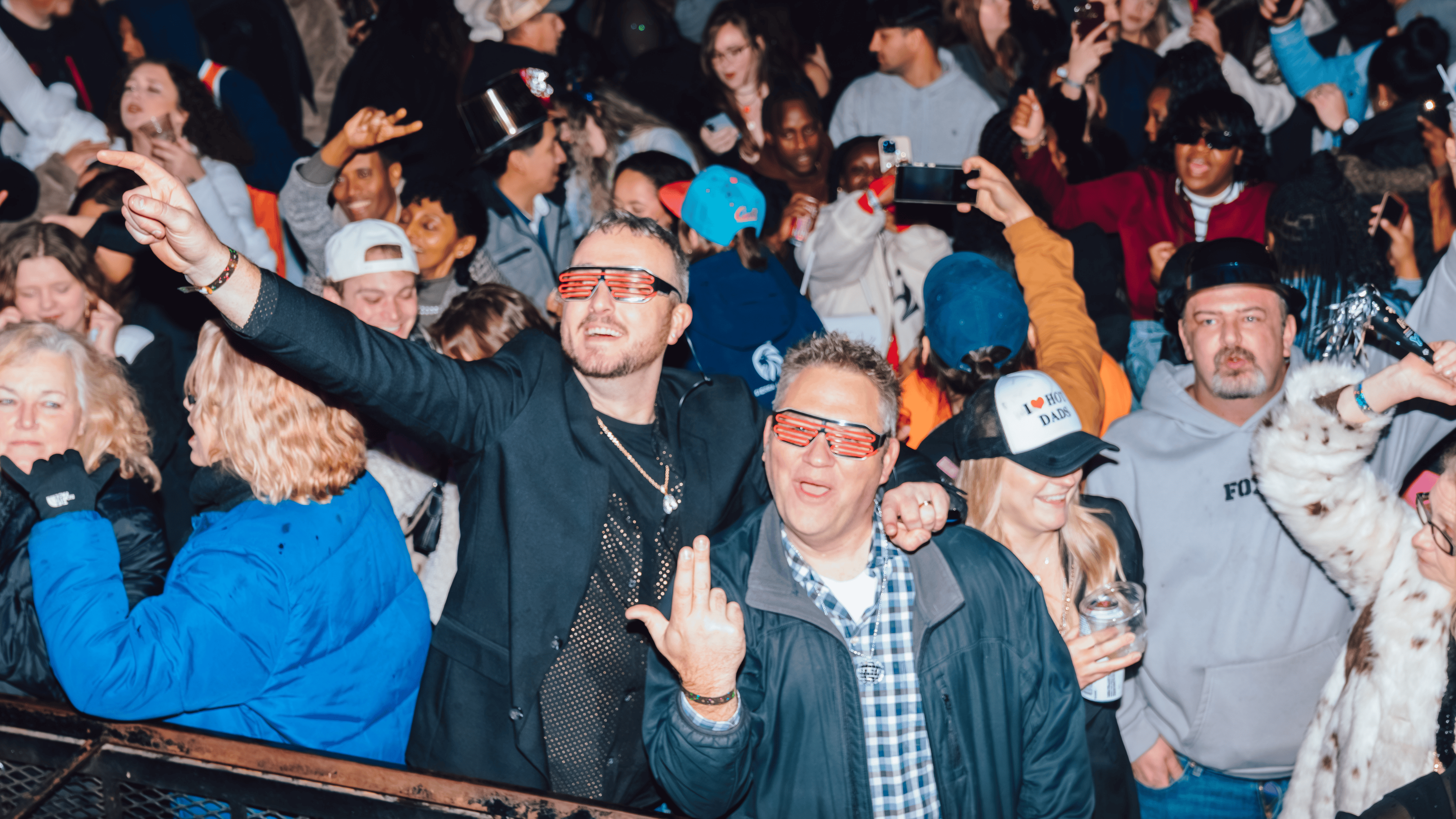 An image of two men in a crowd of people at an NYE party, with one pointing up towards the sky, and the other throwing up an "L" with his hand, for Louisville.