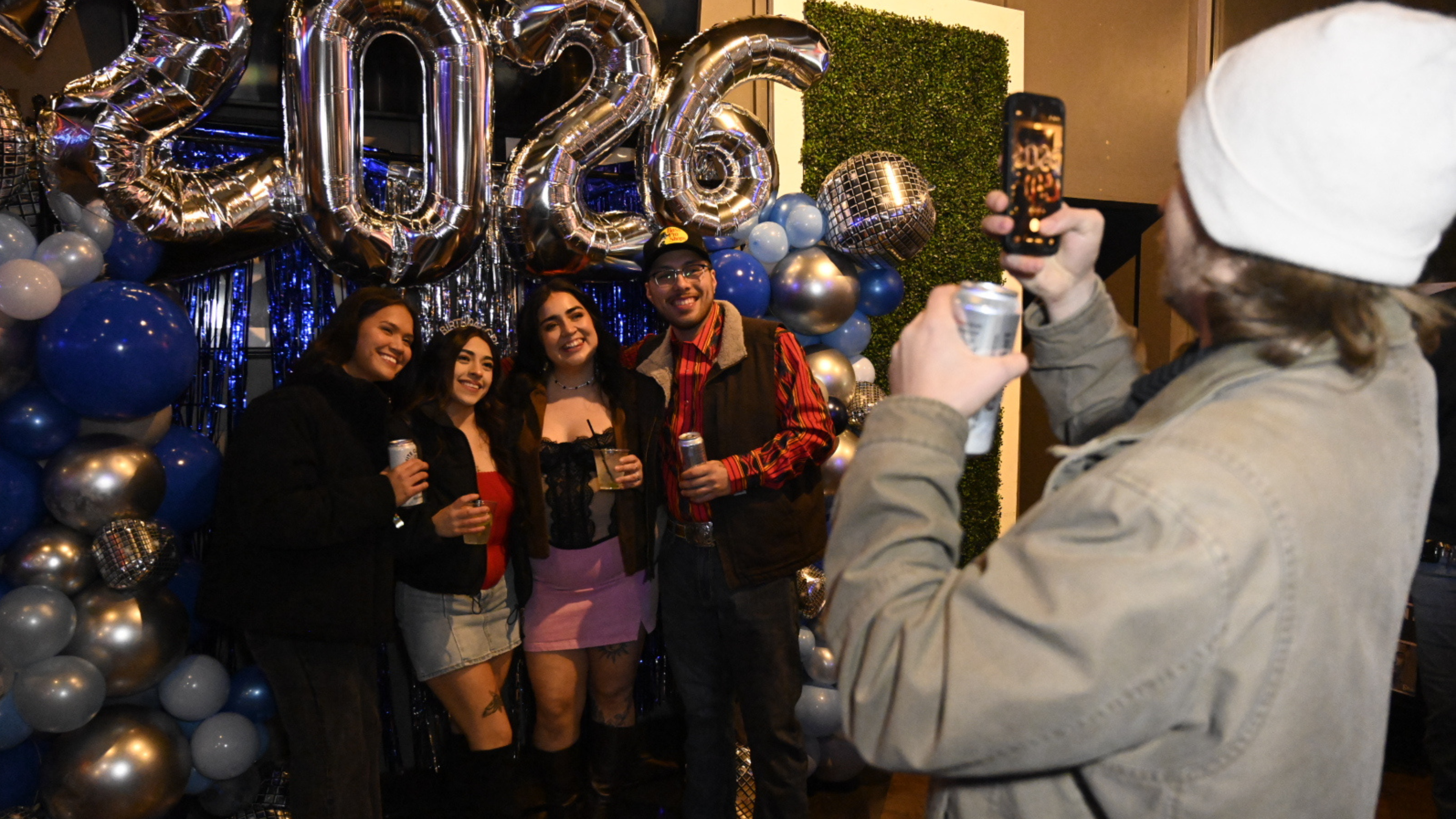 An image of a group of four people posing for a picture in front of a blue, white, and grey backdrop, with grey balloons that say "2026", and a woman taking their picture on a phone.
