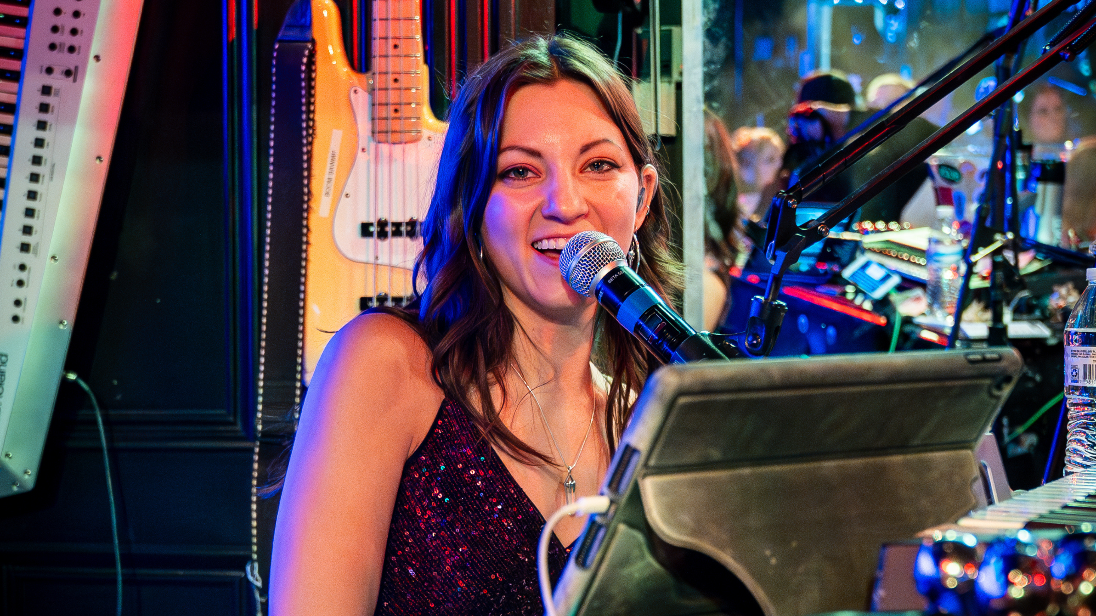 An image of a woman sitting at a piano, singing into the mic, and looking into the camera,.