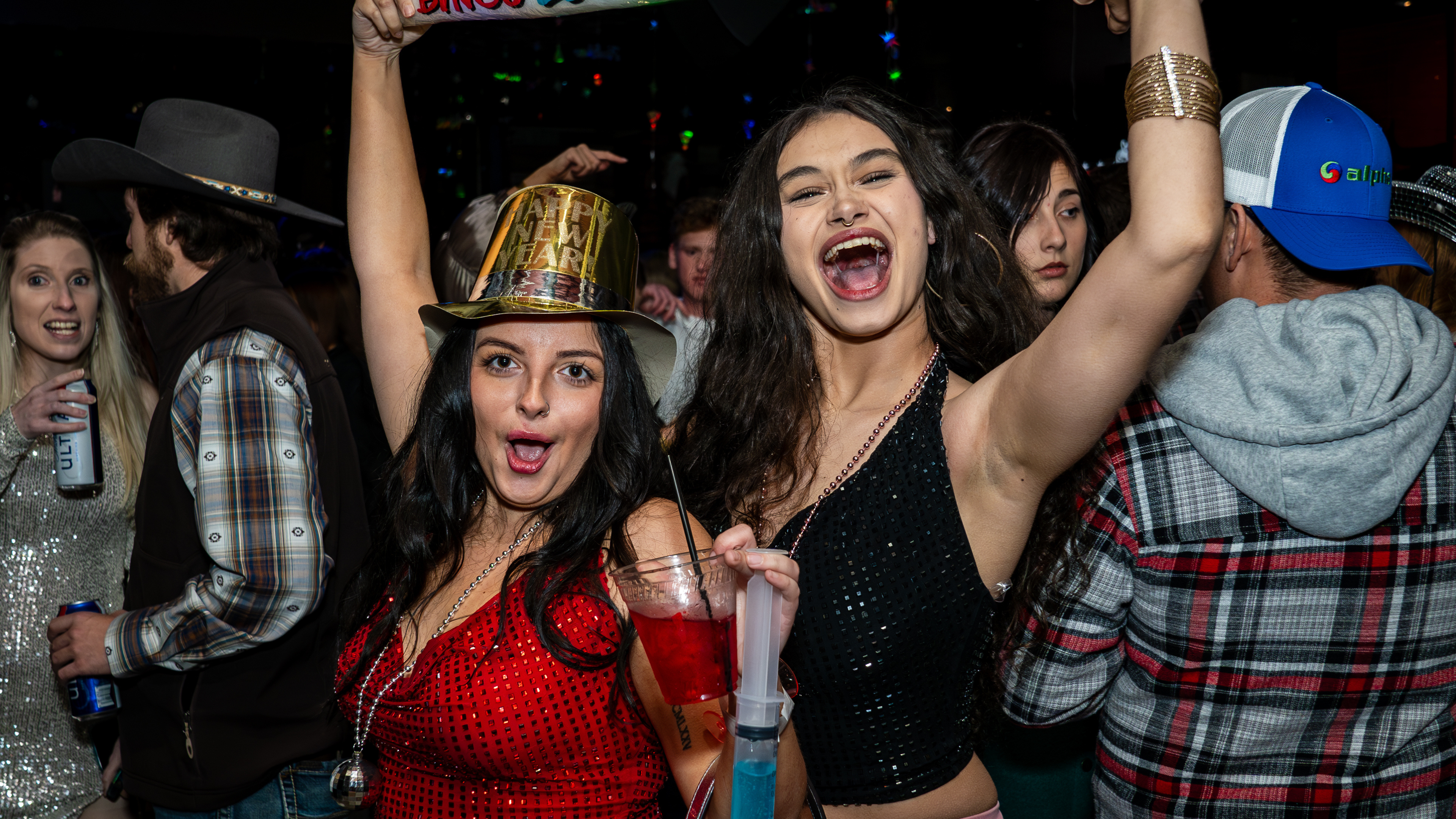 An image of two woman yelling and posing for a picture, one wearing a red top and gold NYE top hat, and the other wearing a black top.