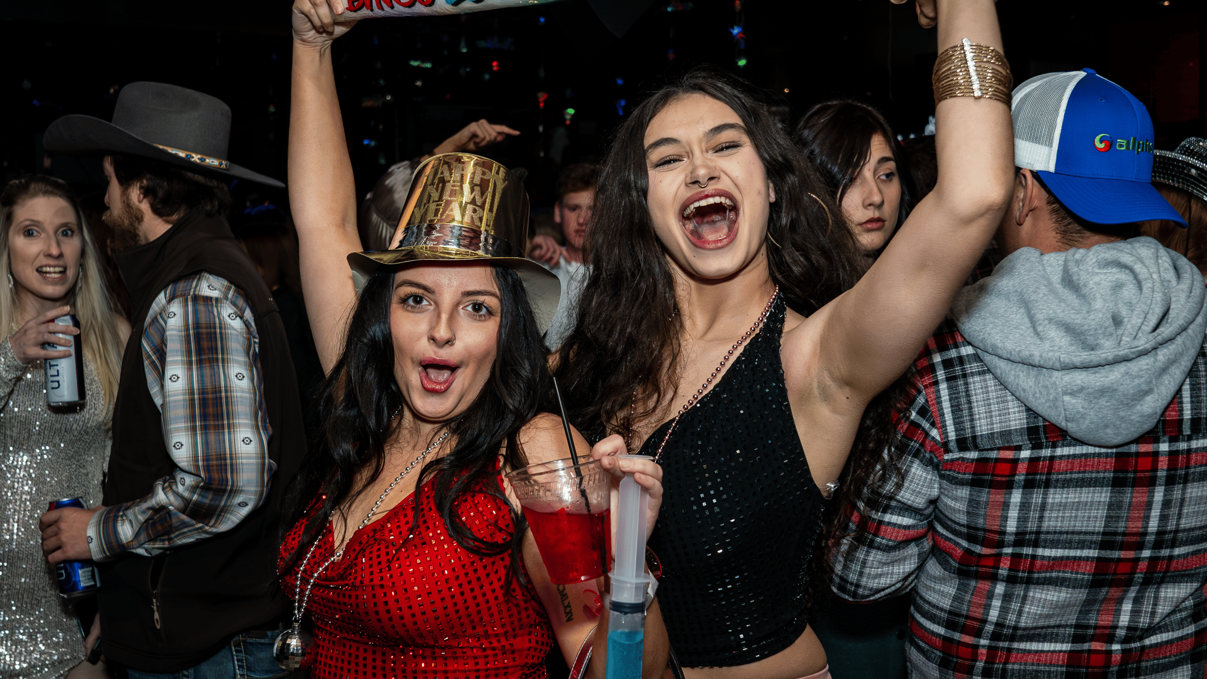 An image of two woman yelling and posing for a picture, one wearing a red top and gold NYE top hat, and the other wearing a black top.