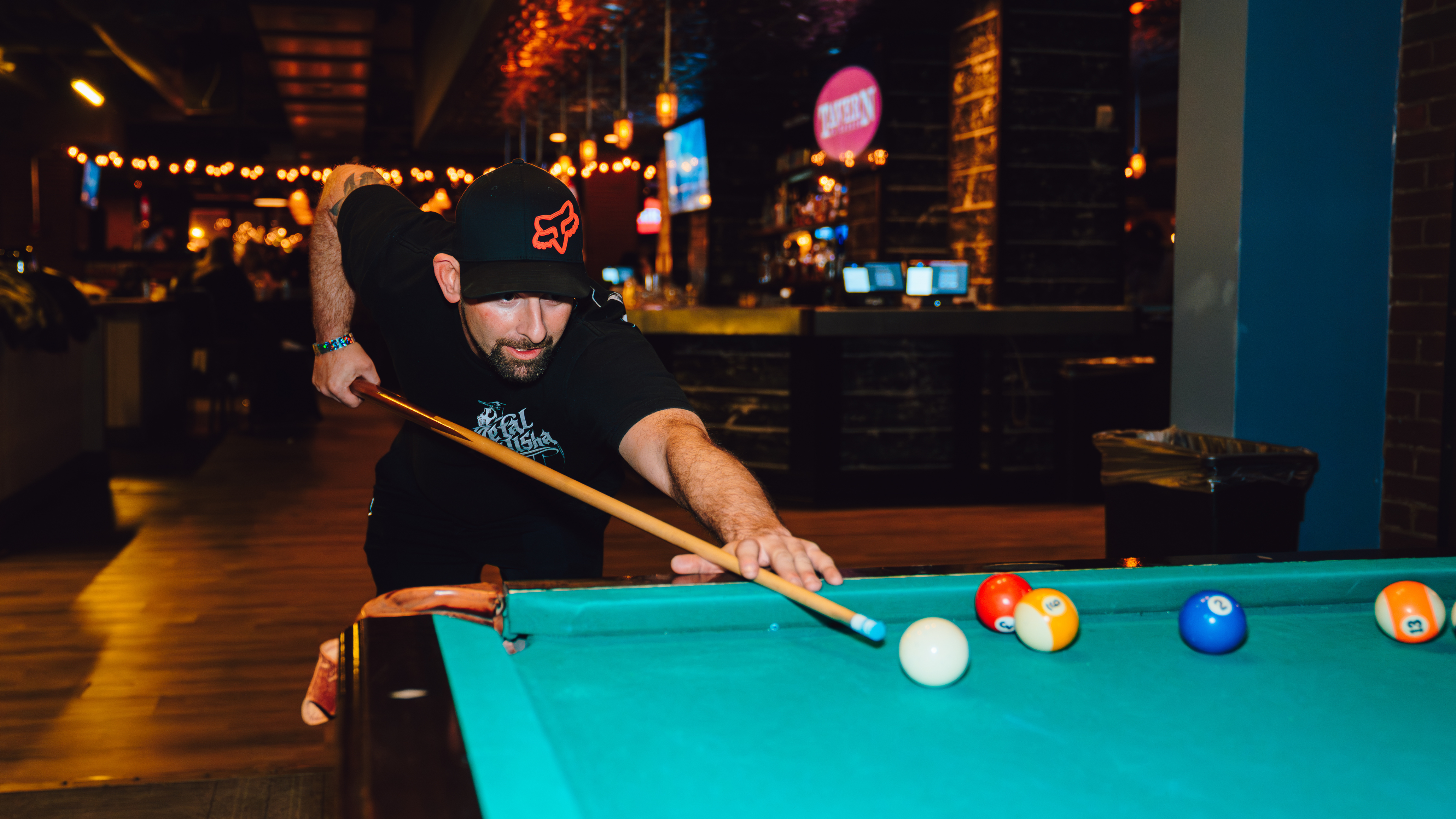 A lower angle image of a man playing pool and wearing a. black hat and shirt.