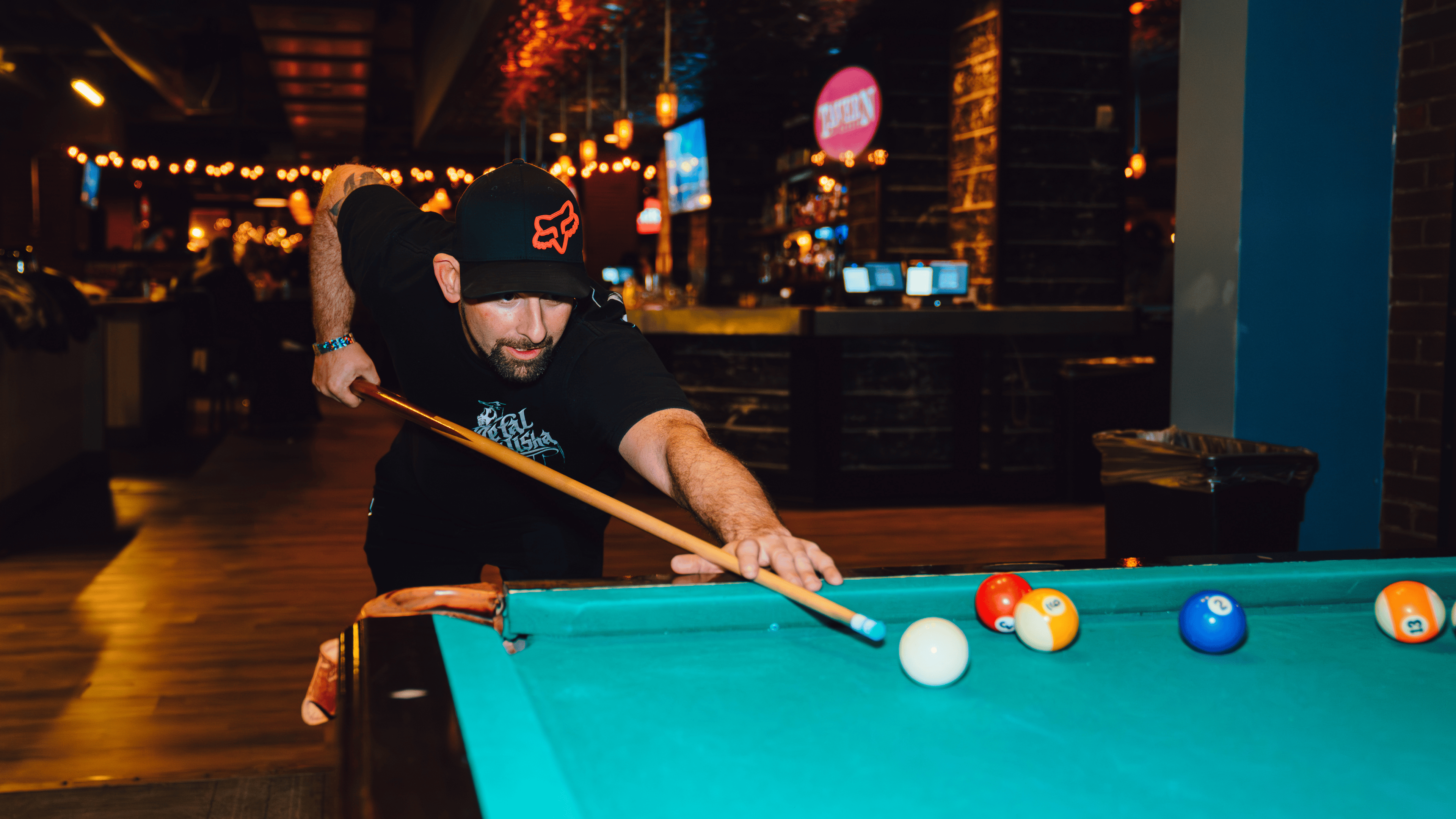 A lower angle image of a man playing pool and wearing a. black hat and shirt.