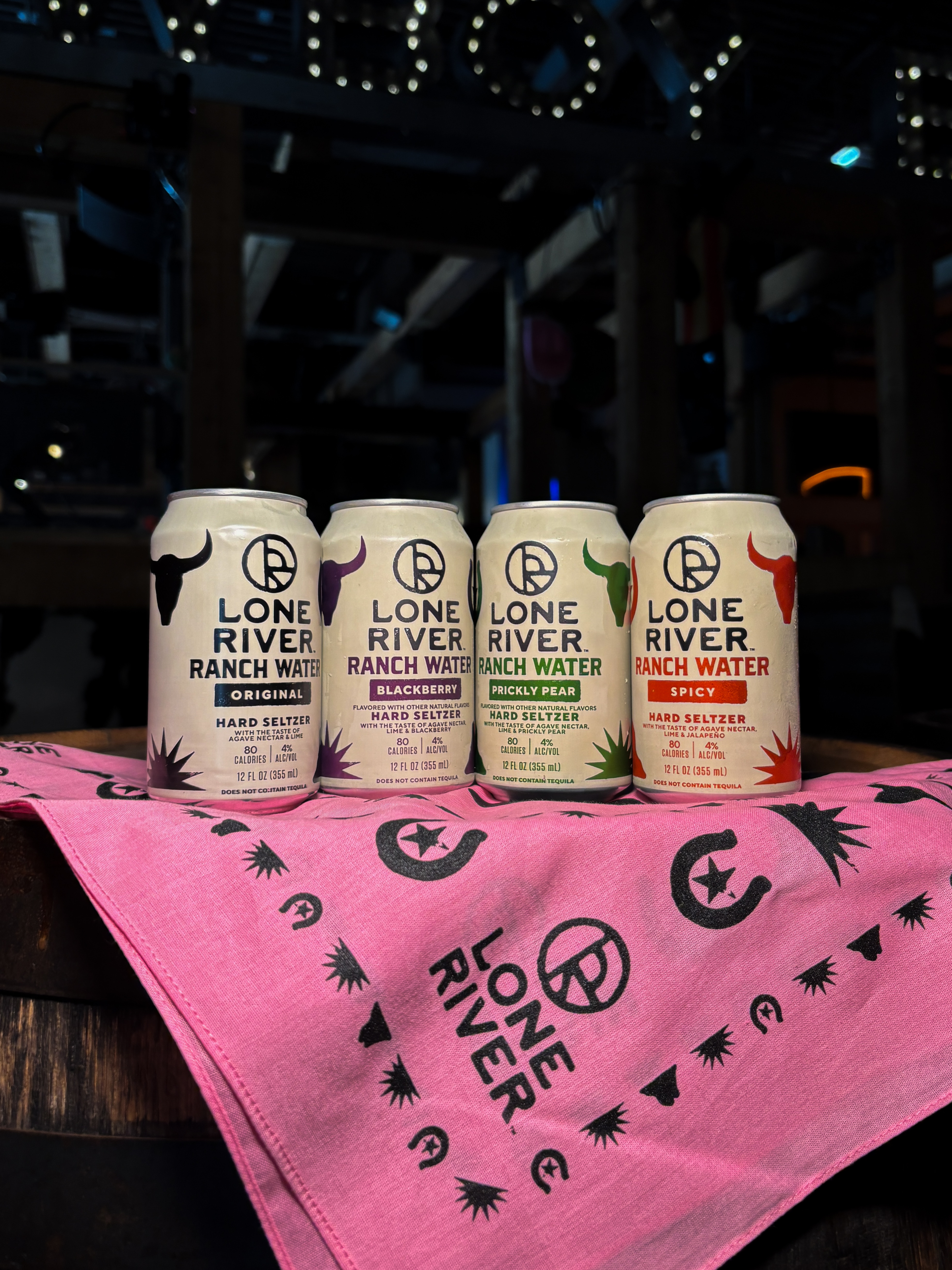 Four (4) Lone River Ranch Waters in our available flavors (original, blackberry, prickly pear, spicy) on top of a pink Lone River bandana containing horseshoe and western imagery.