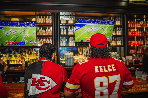 Two men wearing Kansas City Chiefs merchandise facing TV screen watching football