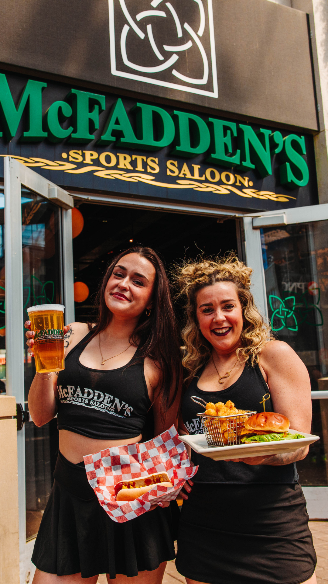 Two bartenders outside of McFadden's holding a burger and fries, a hot dog, and a pint of beer