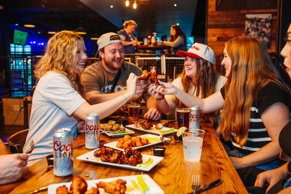 Four people sitting at a table cheering buffalo wings with plates of buffalo wings on the table