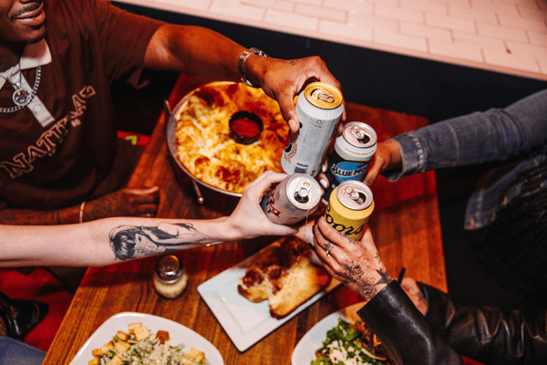 Four hands cheering cans of beer at a table