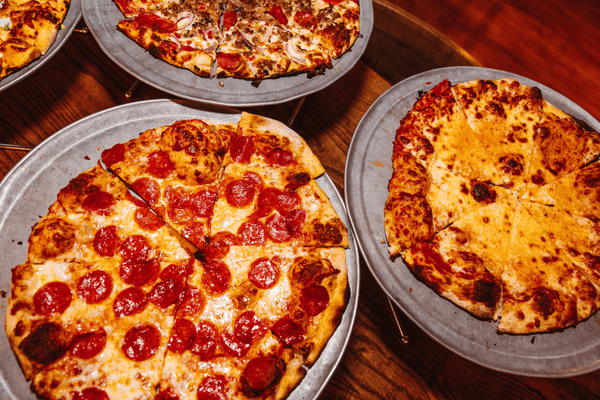 Pepperoni, cheese, and supreme pizzas on metal trays on a wood table