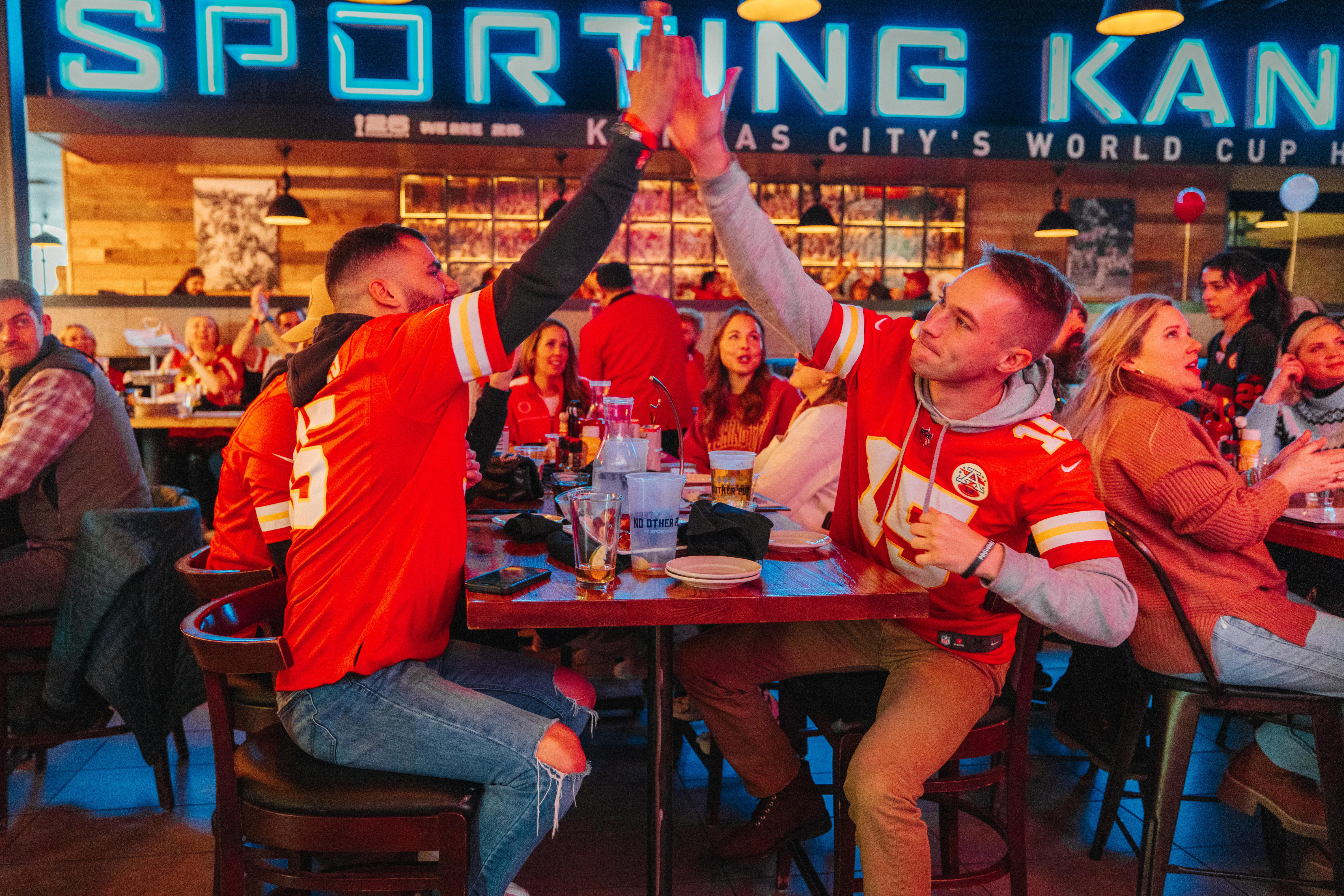 Fans wearing Kansas City football gear high-fiving while sitting at a table