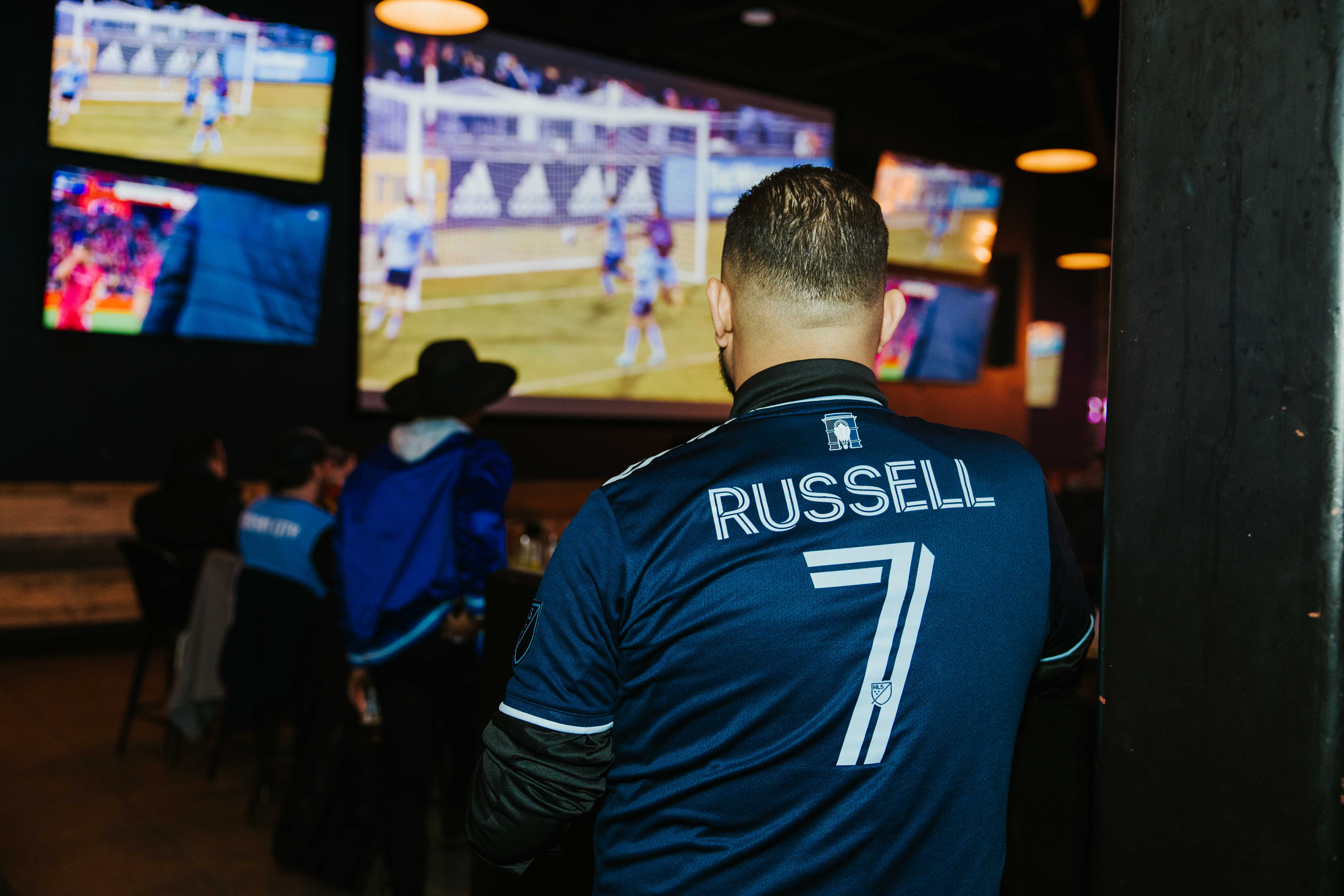 Man wearing soccer jersey watching a match on a big TV screen