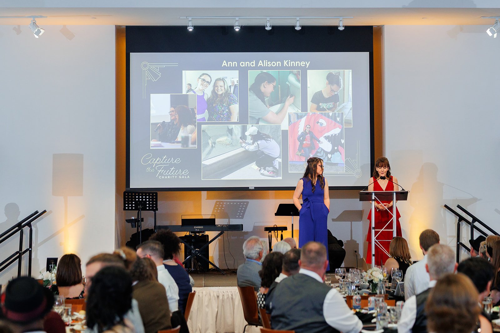 Two people speak on stage to a crowd of guests at The Gallery with a projector in the background.