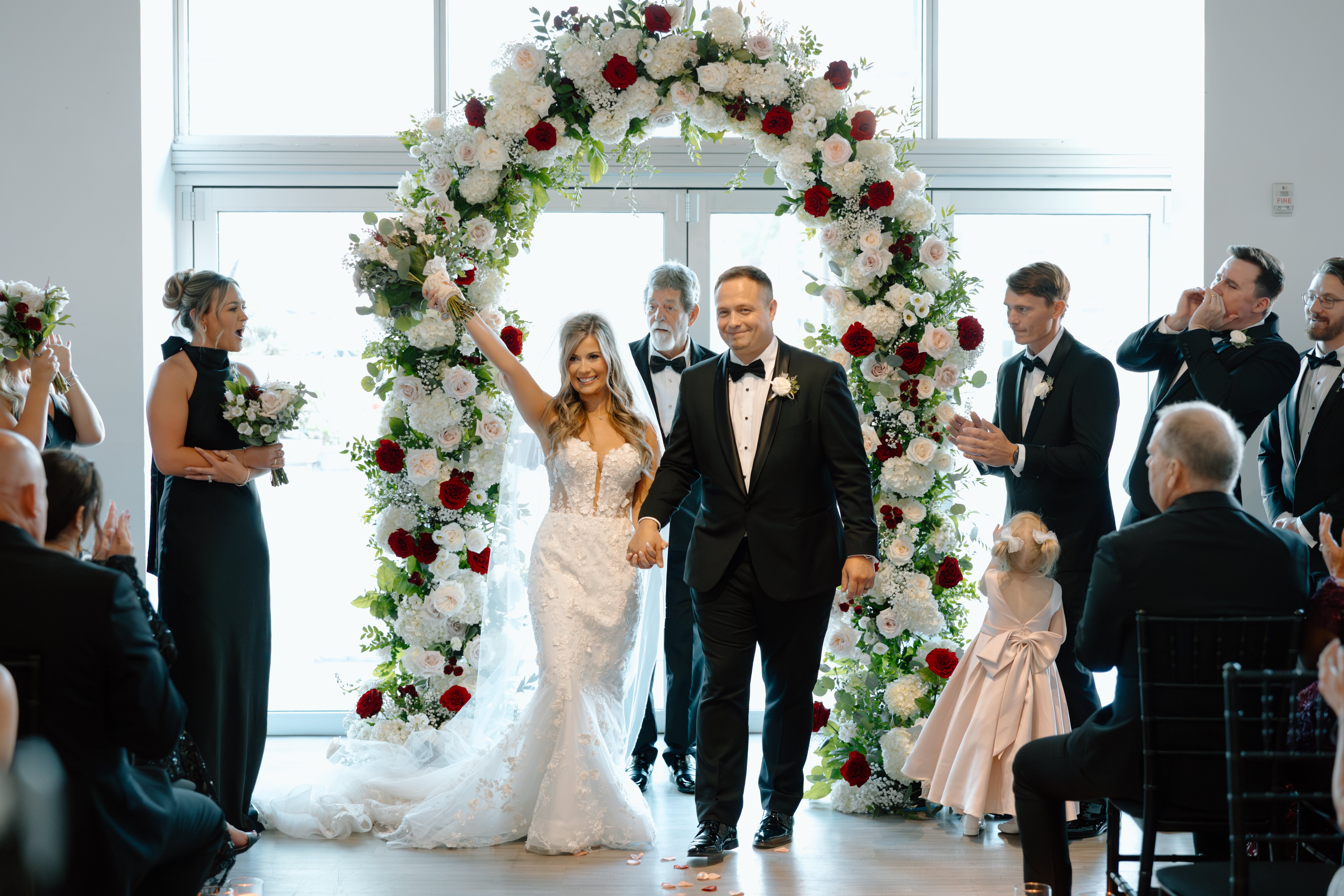 Bride and groom at The Gallery stand in front of a large green and red floral arch.
