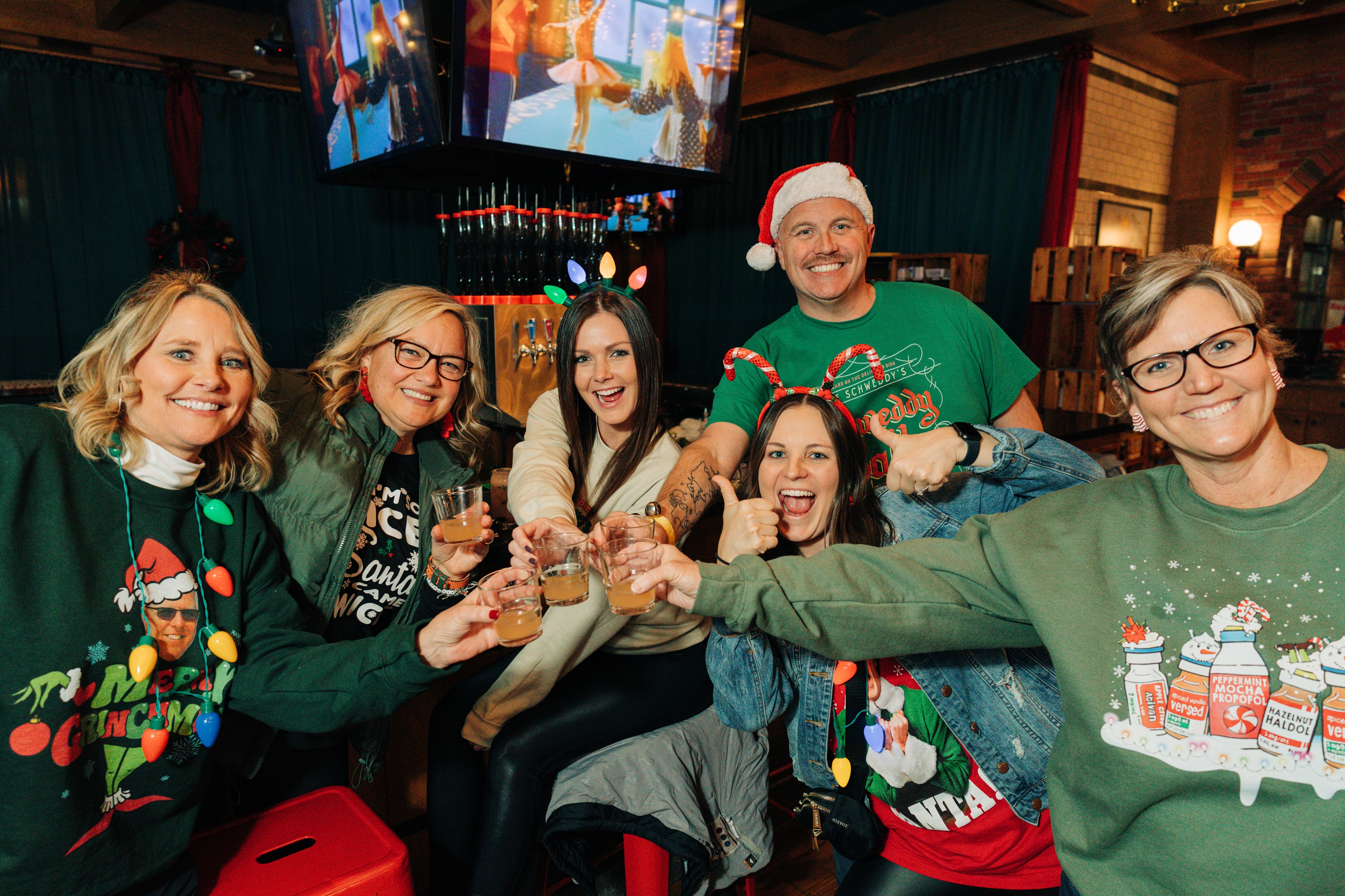 Five women and one man in holiday sweaters and attire toasting cocktails.