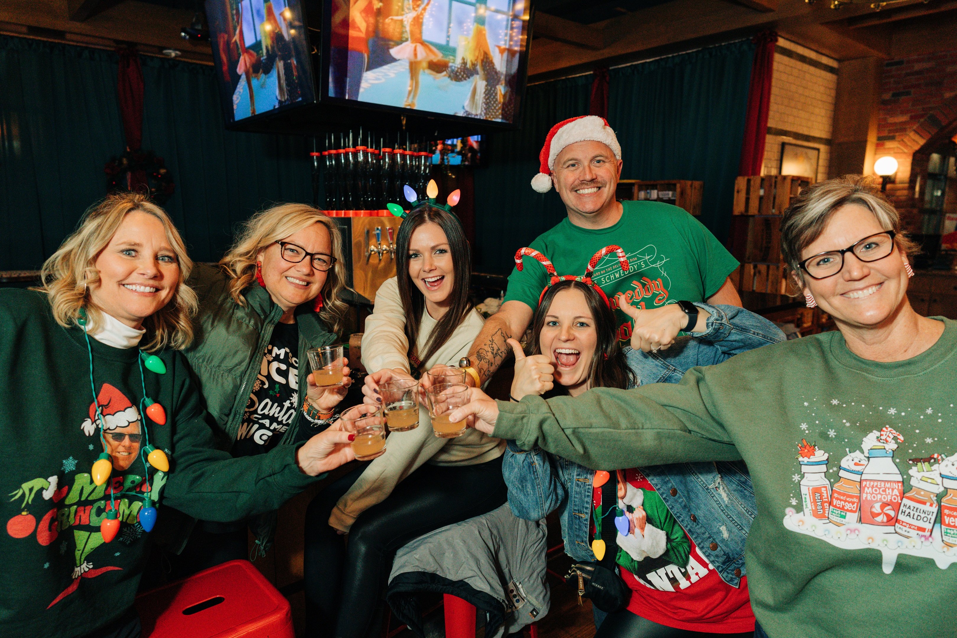 Five women and one man in holiday sweaters and attire toasting cocktails.