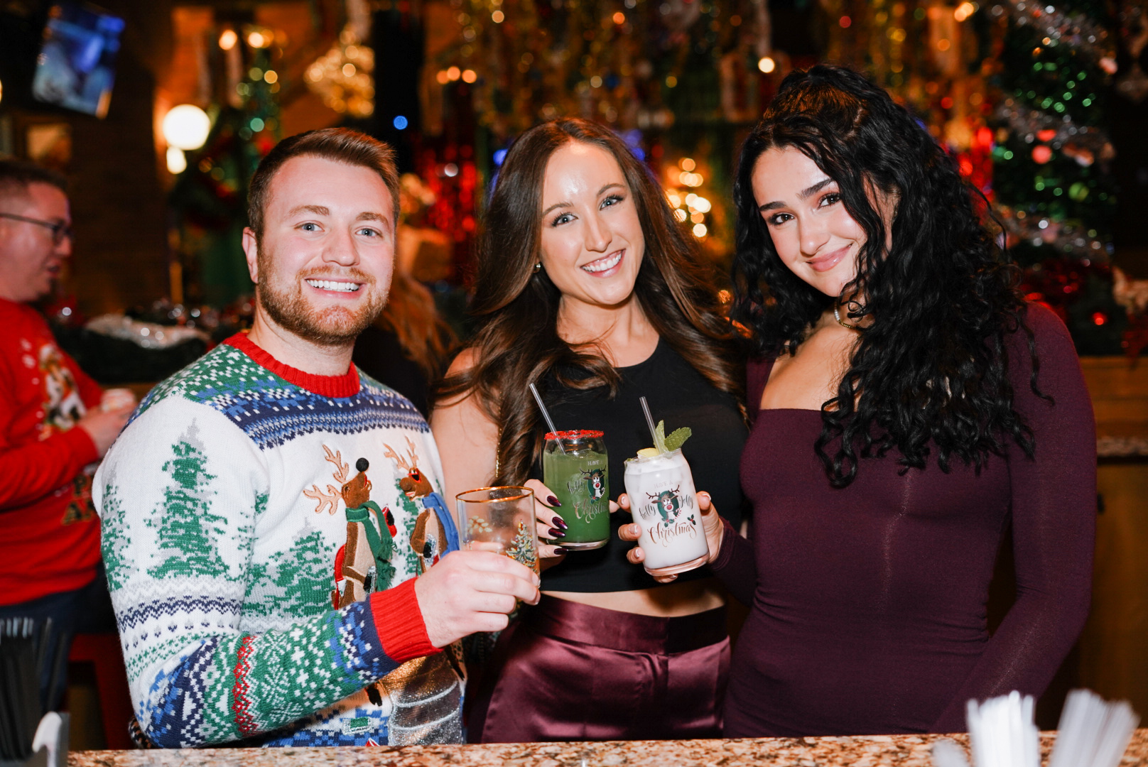 Two women and a man in holiday attire posing with drinks.