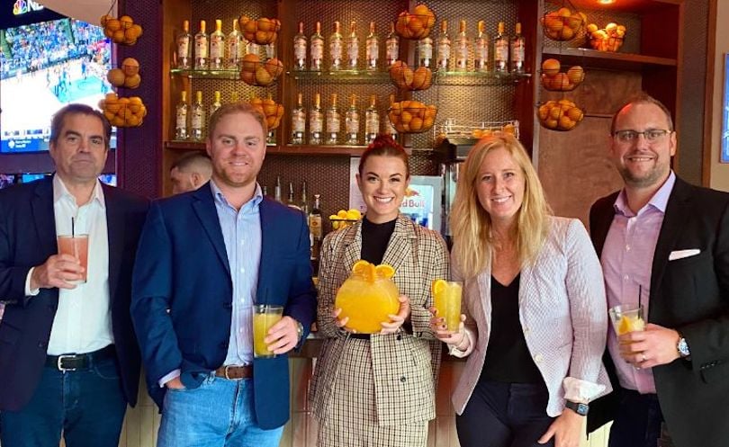 Standing in front of a bar, four smiling leaders surround a fifth who holds a fishbowl filled with a bright orange beverage.