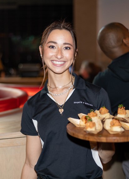Sports & Social female waitstaff carrying wooden tray of food.