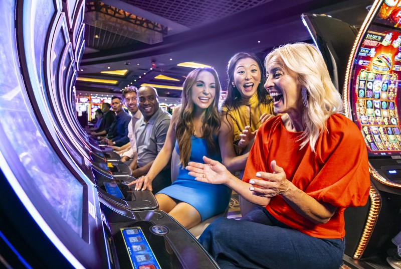 Excited woman in a red blouse and denim slacks sitting at a slot machine with two other women congratulating her win.