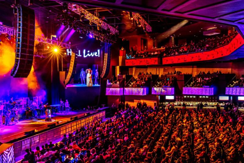 A multi-piece band plays behind a singer on a beautiful purple and gold-lit stage inside a full three-story theater. 