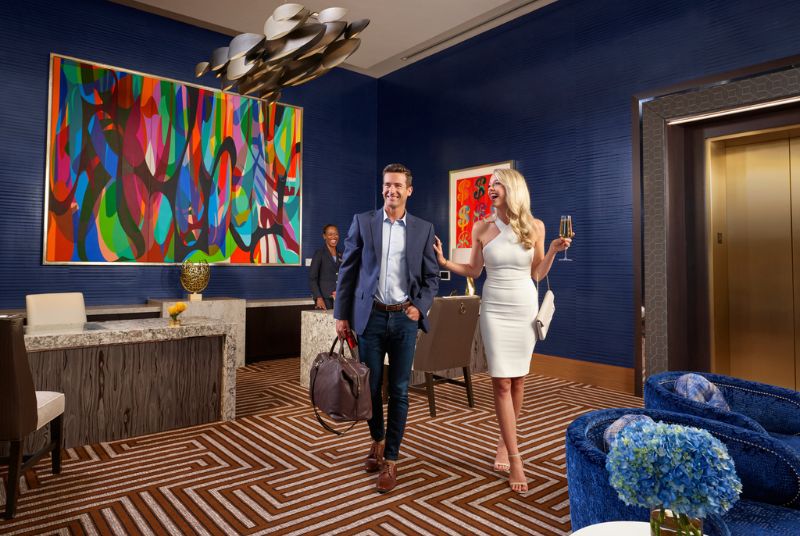 Leaving the VIP desk, a handsome man in a blazer walks with his blonde companion in a white cocktail dress who has champagne.