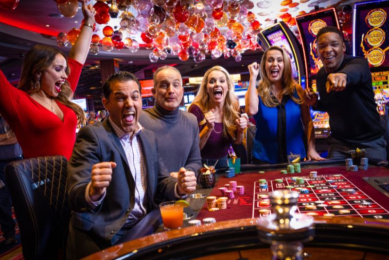 A tableful of excited gamblers cheer and point as a man wins big at roulette, and his companion throws her arms up with joy!