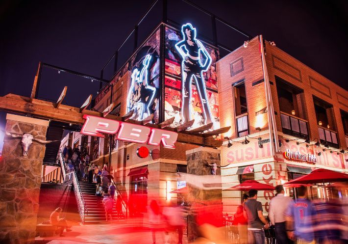 A two-story view of a PBR Cowboy Bar from the outside at night.  People going up the stairs to enter the building.