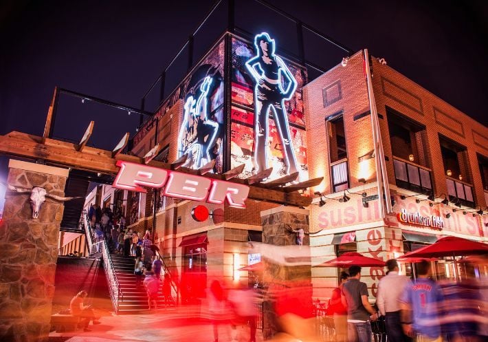 A two-story view of a PBR Cowboy Bar from the outside at night. People going up the stairs to enter the building.