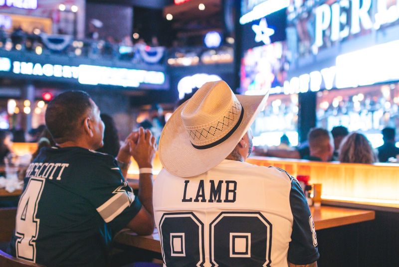 Two Cowboys fans wearing Prescott and Lamb shirts watch as their team plays on the Texas Live! 100-foot LED screen.