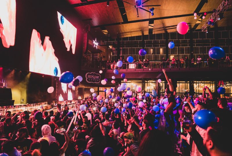  Balloons come down on an enthusiastic crowd as a screen shows blue-eyed fish-like creatures and a DJ is just out of frame.