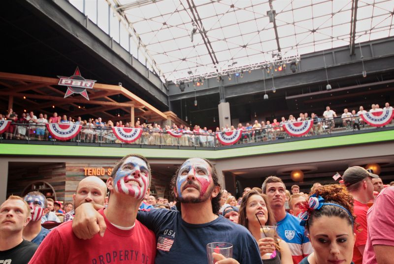 Three men with red, white and blue-painted faces watch USA soccer surrounded by a room full of other enthusiastic fans!