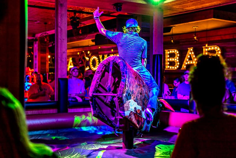 With a backwards baseball hat and an arm raised in the air, a mechanical bull rider takes his turn as the crowd watches!