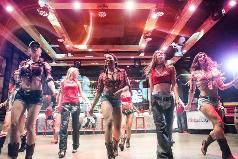 Dressed in tied-off shirts and jean shorts, or tank tops and chaps, a smiling female crew line dances at a PBR Cowboy bar!