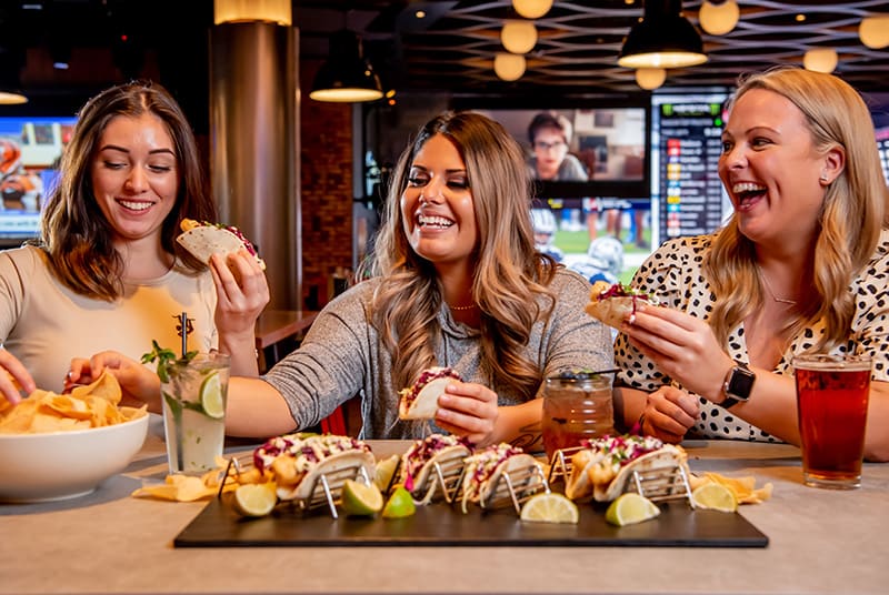 Three happy female friends enjoy beautiful cocktails, tacos, and a bowl full of crispy nachos at Sports & Social!